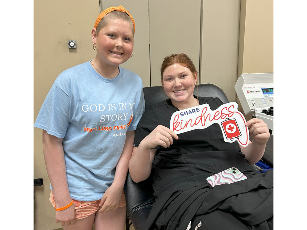 Kaemey Young standing next to a blood donor sitting in chair and holding a share kindness sign.