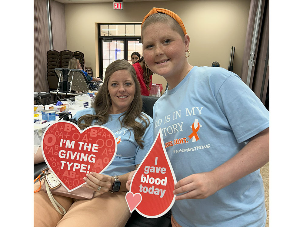Kaemey Young standing next to a blood donor sitting in chair and holding heart shape and blood drop shape blood donation signs.