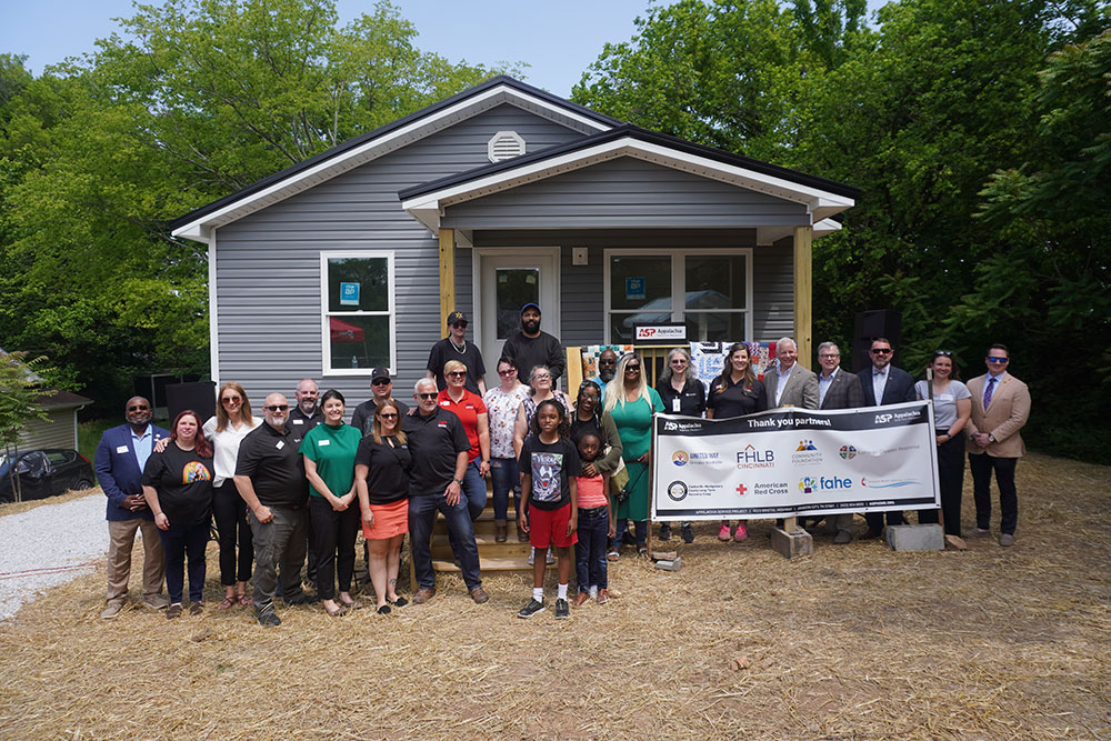 A diverse group of people stands smiling in front of a small, gray house with a porch, holding a banner. The scene is outdoors, surrounded by trees..