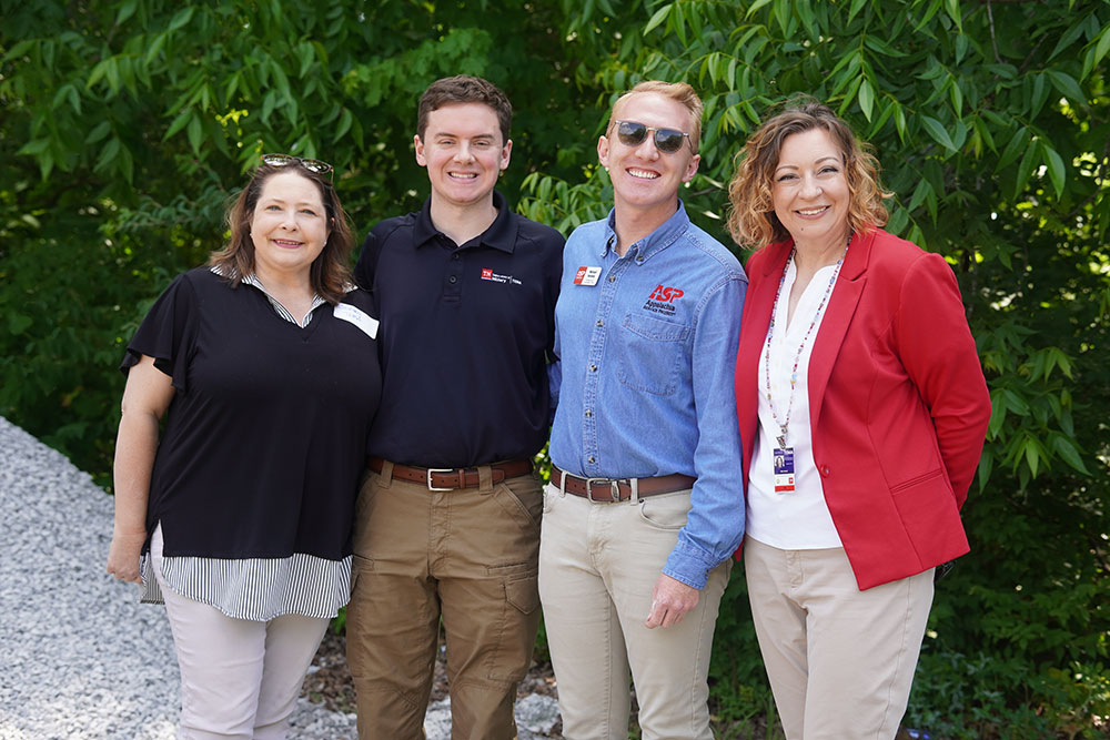 Four smiling people stand together outdoors, surrounded by lush greenery. They are dressed in casual professional attire.
