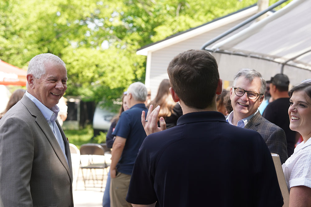 A group of people at an outdoor gathering, engaged in friendly conversation. Everyone is smiling, in a sunny setting with trees and a canopy in the background.