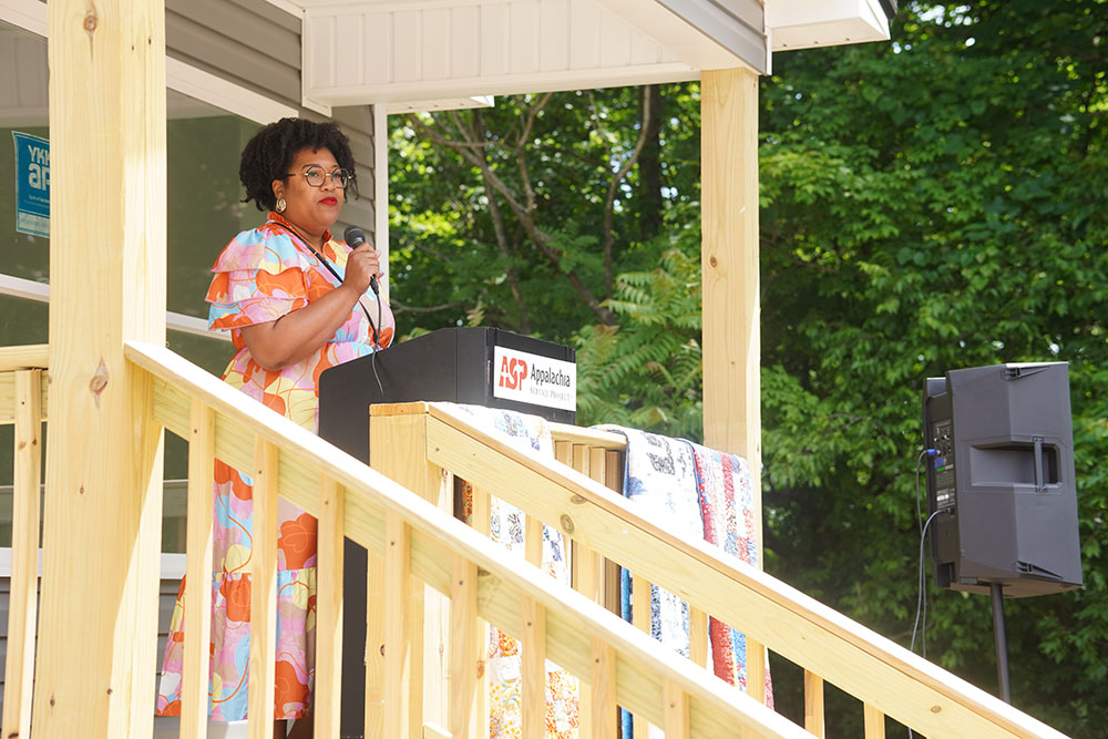 A woman in a colorful dress speaks at a podium on a porch with wooden railings. Lush greenery surrounds the scene.
