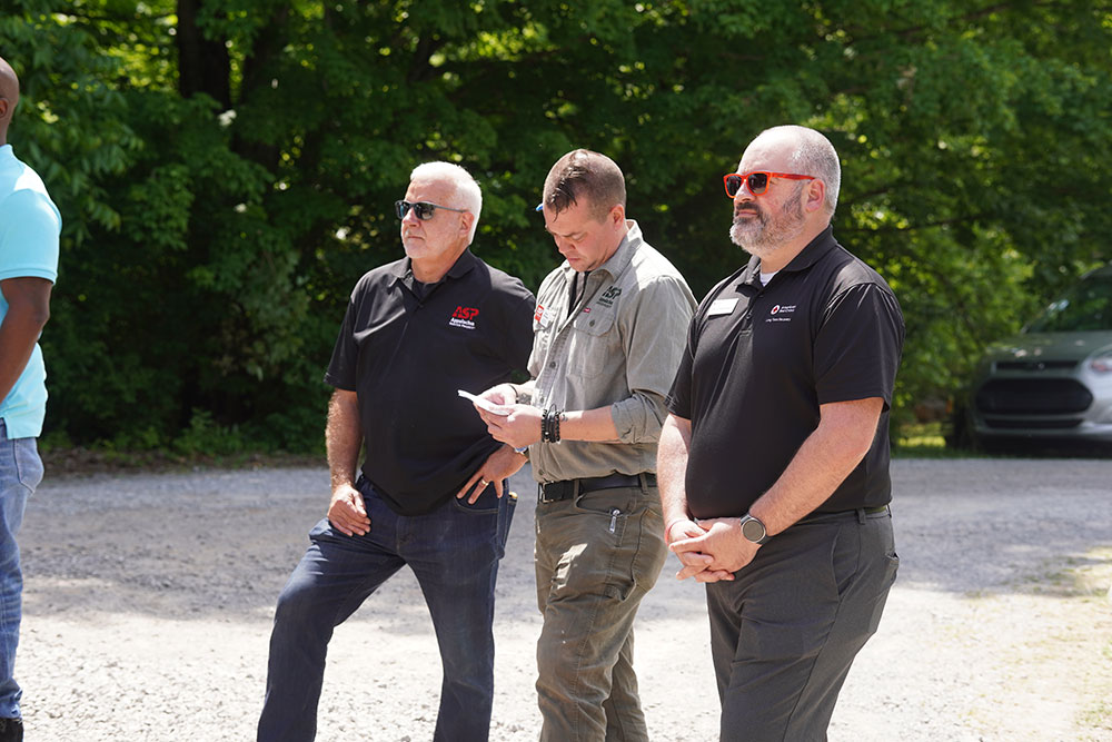 Three people stand outdoors on a gravel path, with trees in the background. They wear casual attire, one writing notes.