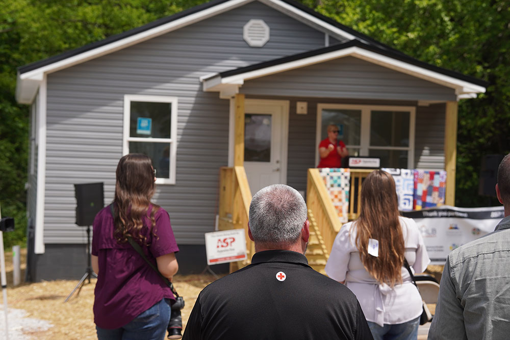 A small gray house with a porch hosts a speaker at a mic. A small audience listens, surrounded by greenery.