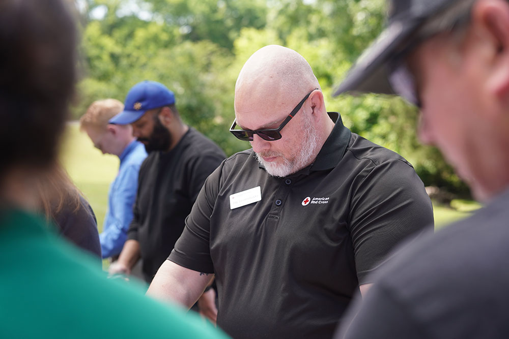 A bald man wearing sunglasses and a black polo with a Red Cross logo stands in a group, heads bowed in a moment of reflection, outdoors in sunlight.
