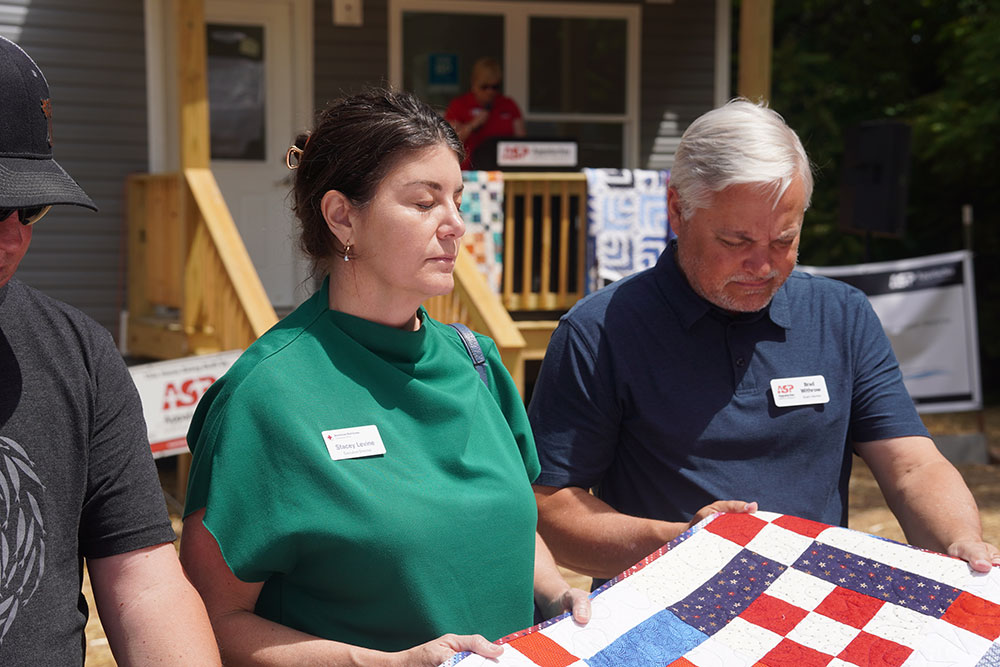 Three people stand solemnly holding a colorful patchwork quilt outside a house. They appear reflective and respectful, with eyes closed.