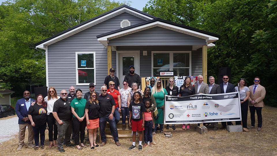 A diverse group of people stands smiling in front of a small, gray house with a porch, holding a banner. The scene is outdoors, surrounded by trees..