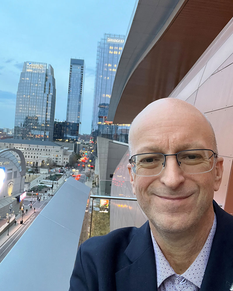 Ralph Campbell, wearing suit and glasses, standing on a balcony of a tall building, takes a selfie.