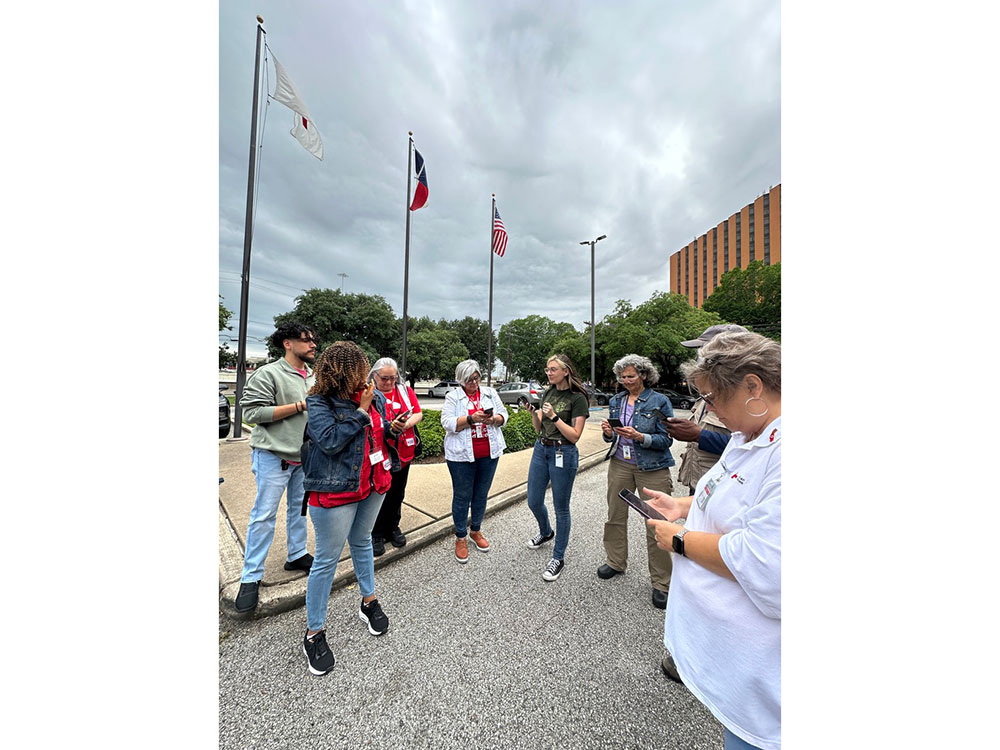 Red Cross volunteers in parking lot next to flag poles looking at cell phones