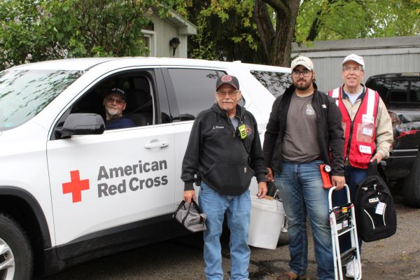 Three Red Cross volunteer standing next to a Red Crosscar with a person in the car.