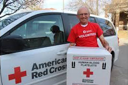 Red Cross volunteer carrying a large box and standing next to a Red Cross vehicle.