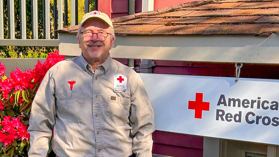 Red Cross nurse volunteer Bill Szabo wearing glasses, hat and Red Cross outfit, smiles for picture.