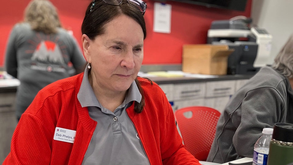 Red Cross nurse volunteer Deb Phelps wearing glasses on top of head and Red Cross name tag while sitting at a desk.