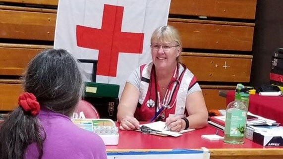 Red Cross nurse volunteer Lynn Jackson wearing glasses, Red Cross outfit and a stethascope around neck, sitting at table with another person across from her.