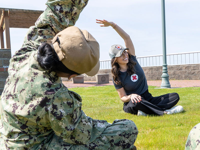 A Red Cross volunteer and a military service member stretching on the grass on a sunny day.