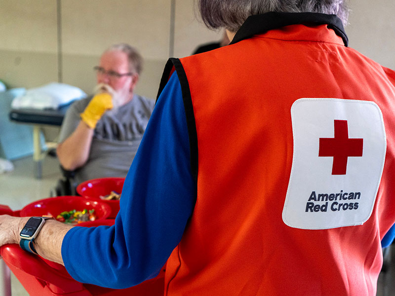 A Red Cross volunteer carrying a tray of snacks and walking towards a military service member.