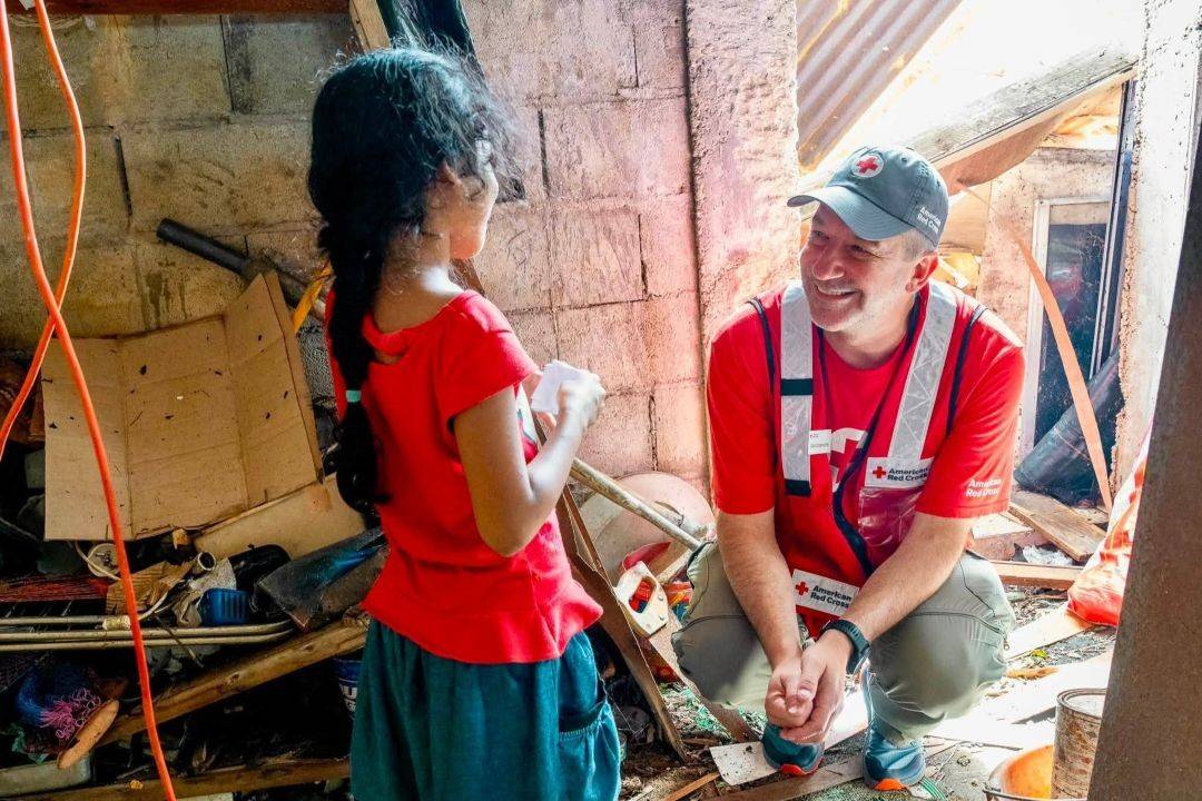 Man in Red Cross shirt and hat smiles at young girl while standing in a home destroyed by disaster