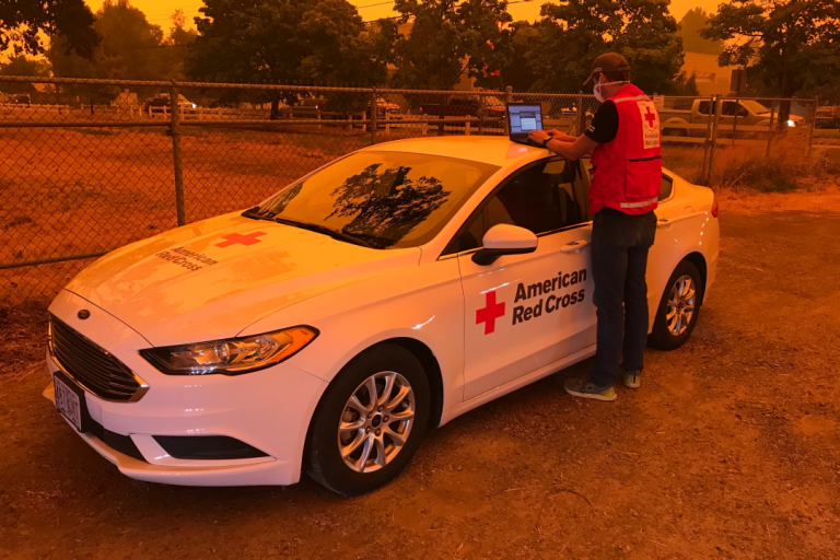 Man works on laptop near Red Cross vehicle with orange glow from wildfires
