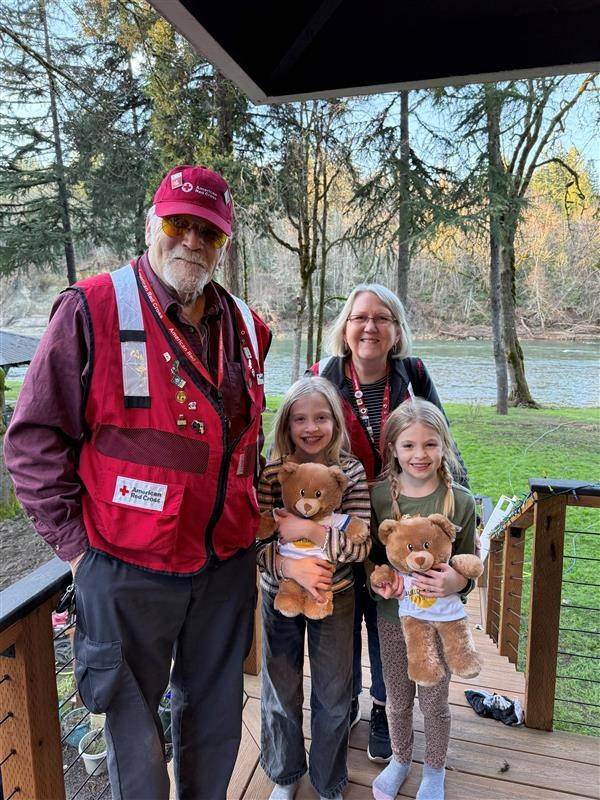 Two Red Cross volunteers stand on porch with young children holding stuffed animals