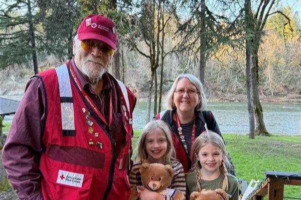 Two Red Cross volunteers stand on porch with young children holding stuffed animals