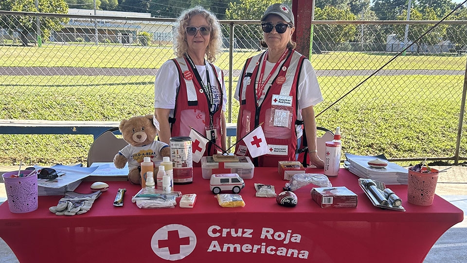 Dos voluntarios de la Cruz Roja junto a una mesa con artículos promocionales de la Cruz Roja.