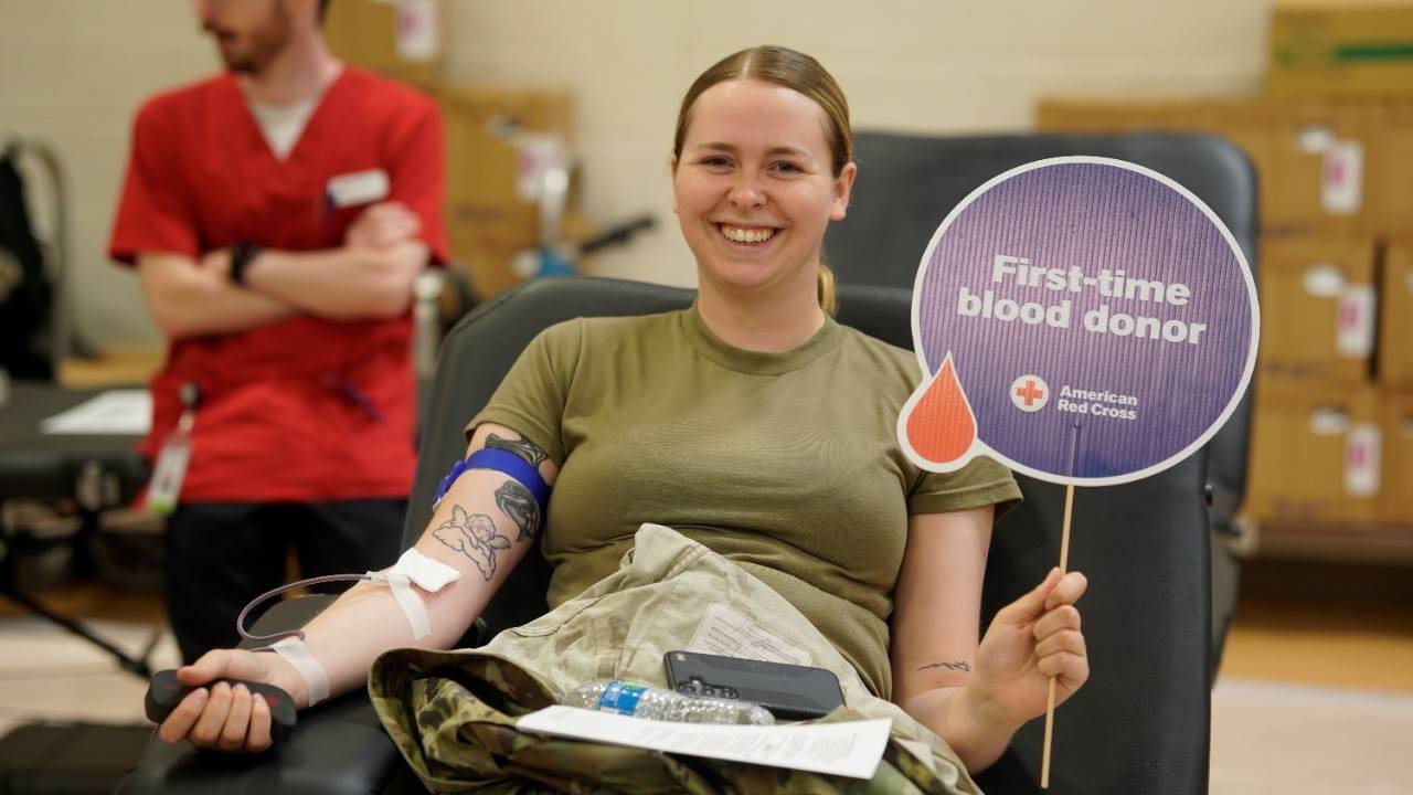Woman in military uniform holds up sign that says "First Time Blood Donor" while donating blood