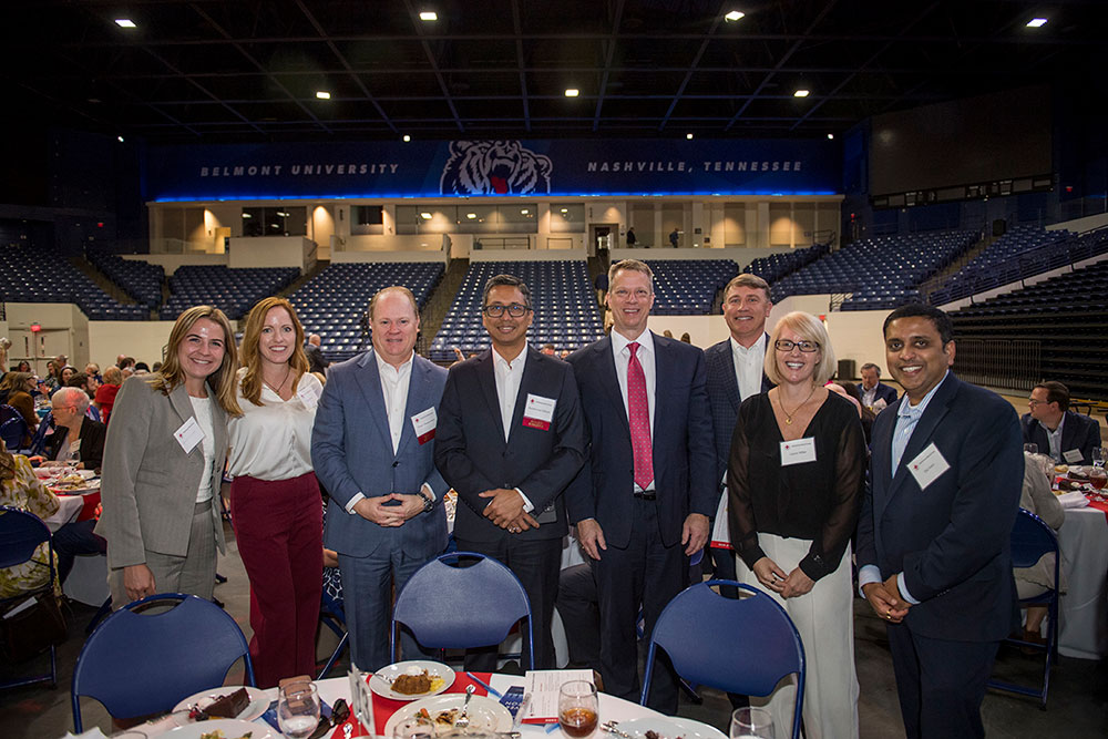 A group of well-dressed individuals smiling at a formal event in a large auditorium. The banner above reads "Belmont University, Nashville, Tennessee."