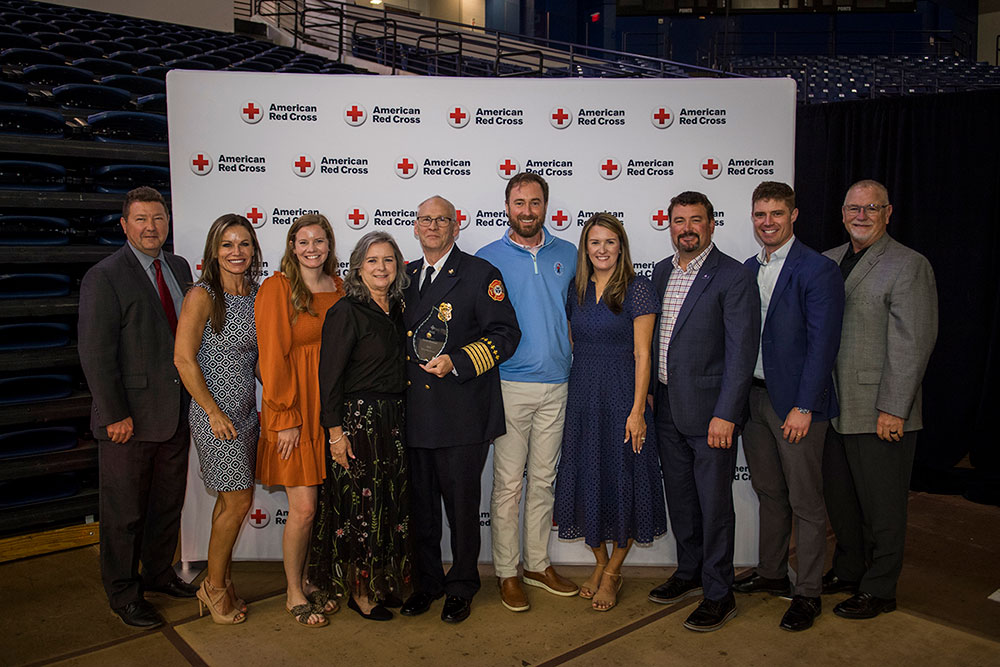 A group of ten people, including a uniformed firefighter holding an award, pose smiling in front of an American Red Cross backdrop in an auditorium.
