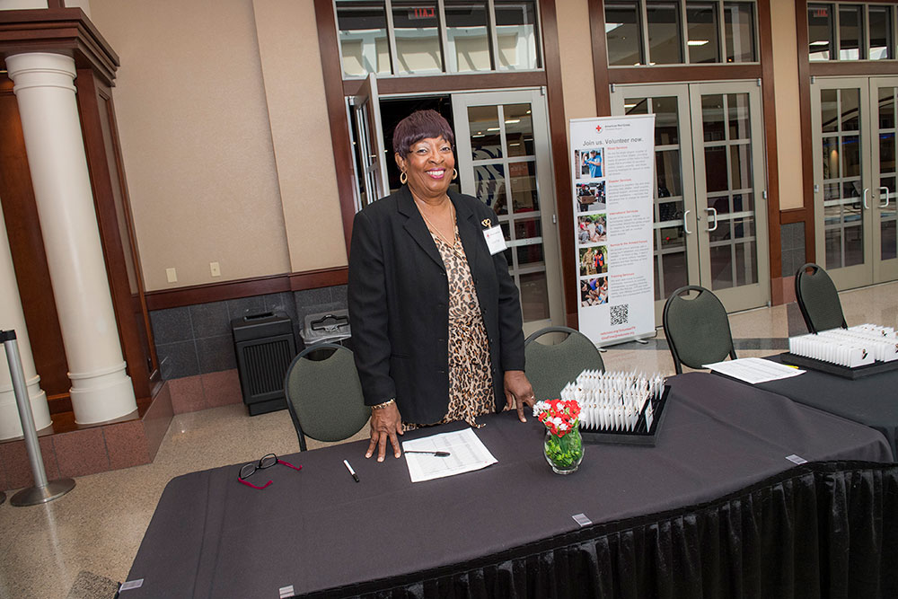An older woman, smiling in a conference setting, stands at a registration table with a sign-up sheet, pens, and flowers. She wears a black blazer and patterned blouse.