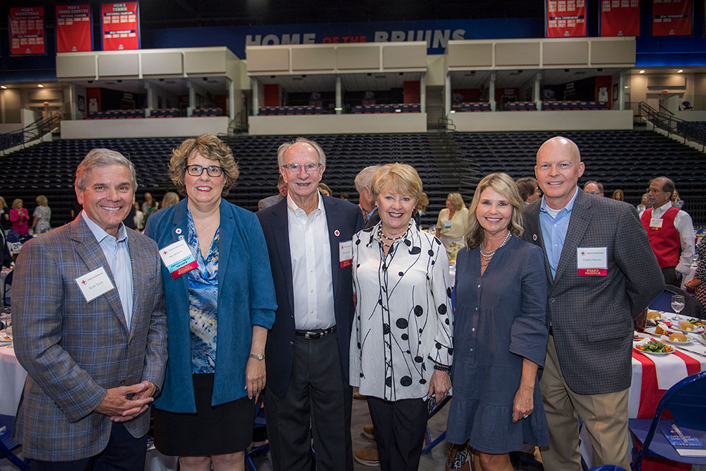 A group of six adults, smiling and posing together in an indoor sports arena. They are dressed in semi-formal attire, under a "Home of the Bruins" sign.