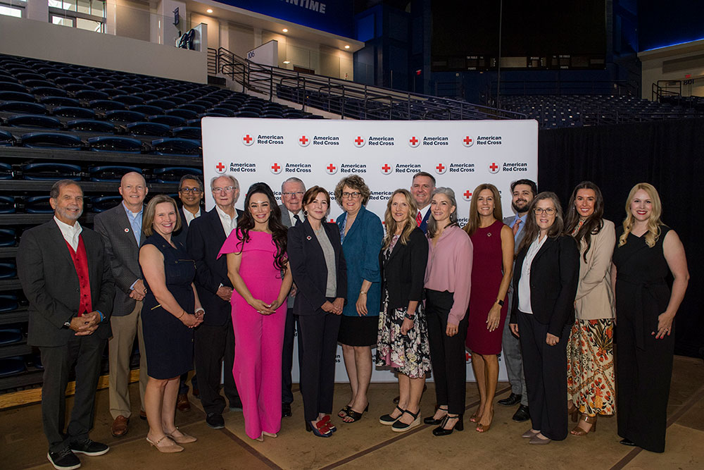 A diverse group of 16 people, dressed formally, stands in front of an American Red Cross banner inside a large auditorium. They appear cheerful and united.