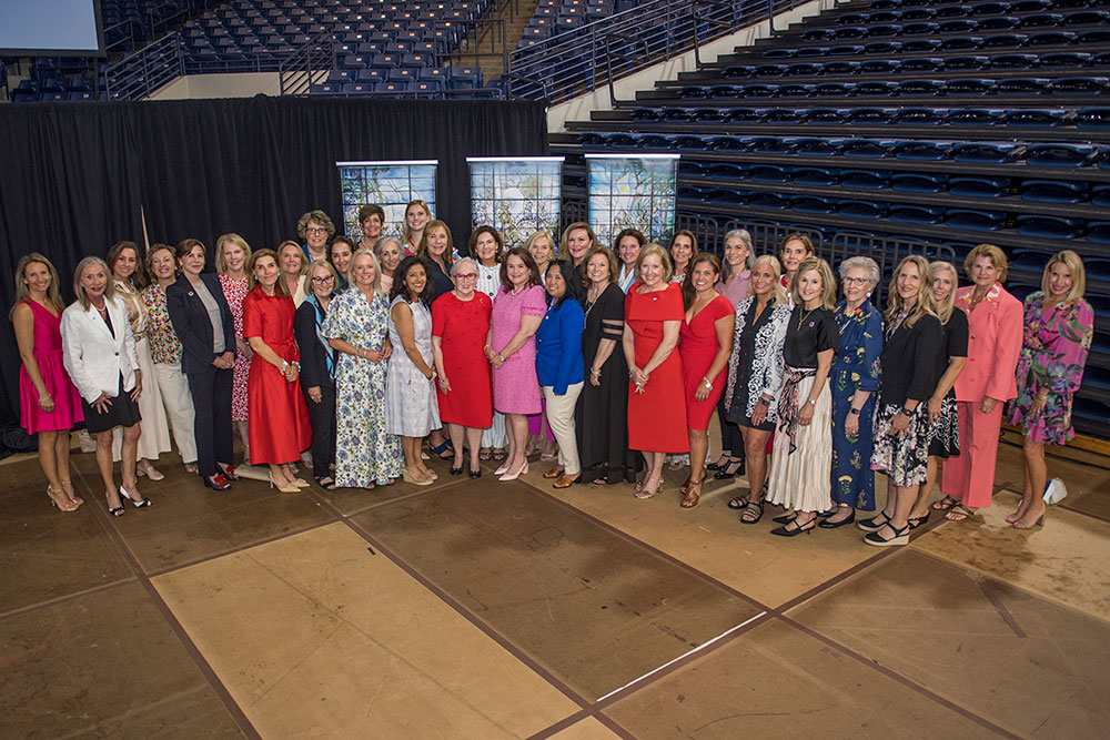 A group of women stand together in a gymnasium, wearing colorful dresses and business attire, smiling warmly at the camera, conveying a sense of unity.