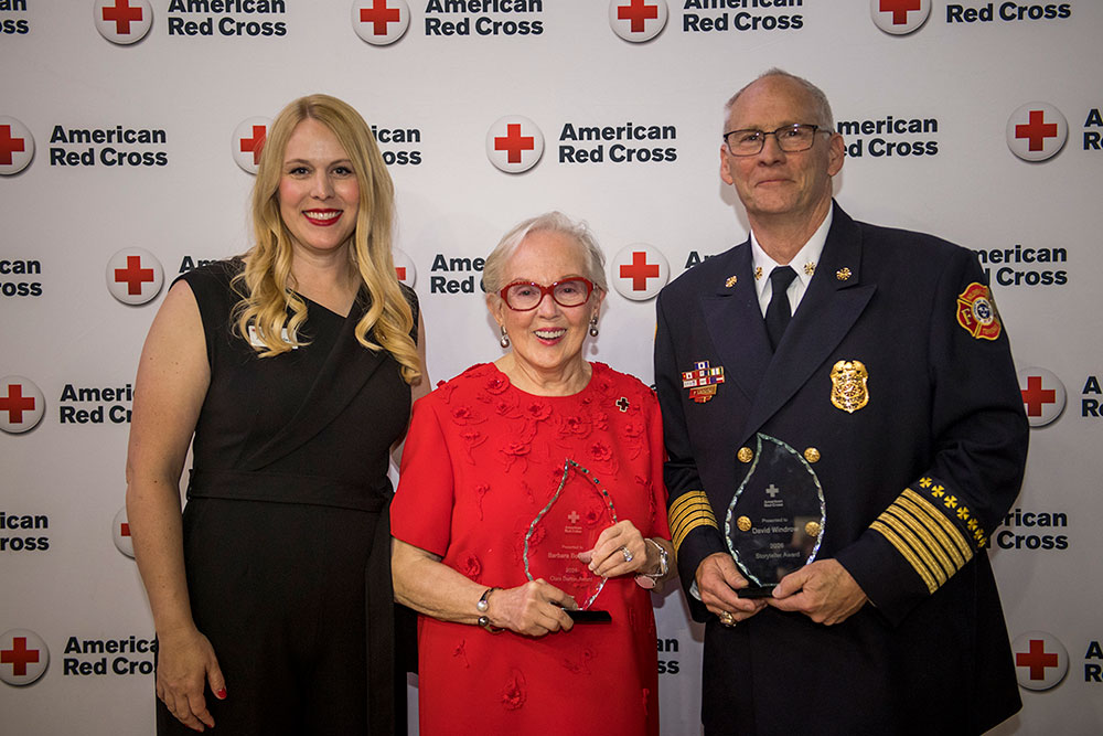 Three people smiling at the American Red Cross event. The center woman holds an award, flanked by another woman and a man in a uniform.