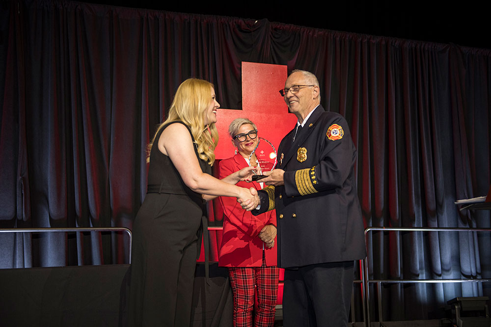 A woman in a black dress smiles as she receives an award from a uniformed firefighter. A woman in red plaid stands smiling in the background.