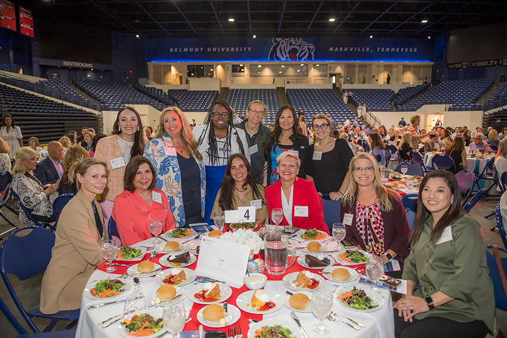 A group of smiling people at a round banquet table with plates of salad and bread rolls in an indoor arena, under a Belmont University sign.