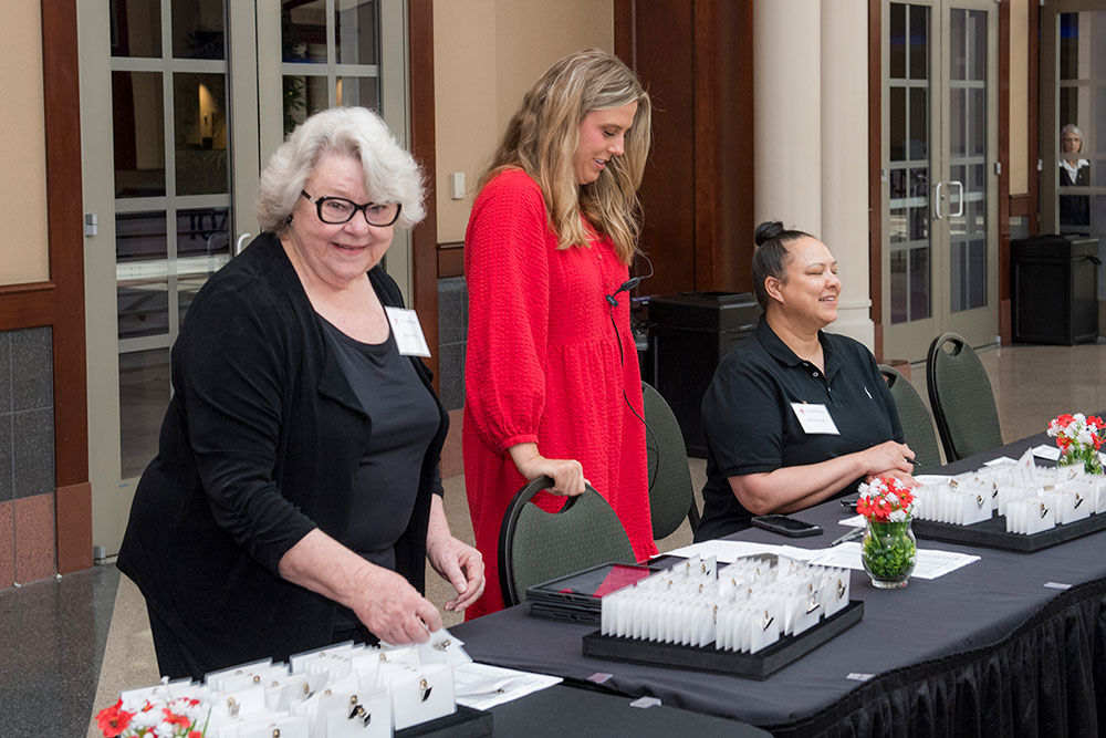 Three women smile and socialize behind a registration table covered with name tags and small red flower arrangements, creating a welcoming atmosphere.