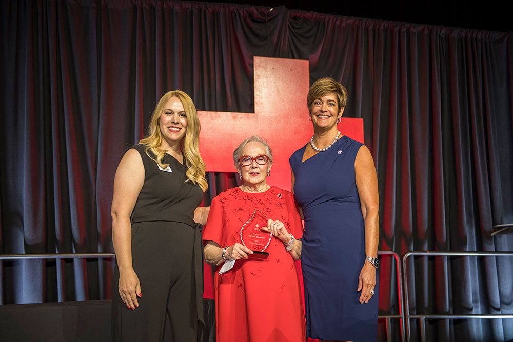 Three women stand smiling against a red cross backdrop. The woman in the center, holding an award, wears a red dress, flanked by two women in black and blue dresses.