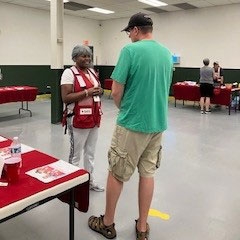 room with folding tables with Red Cross items on them and a Red Cross volunteer speaking to a person.