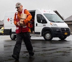 Red Crosser carries supplies while walking to the left, away from an emergency response vehicle with an open door