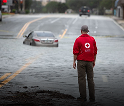 a Red Cross volunteer stands looking at flood waters on a road with a partially submerged car in the distance