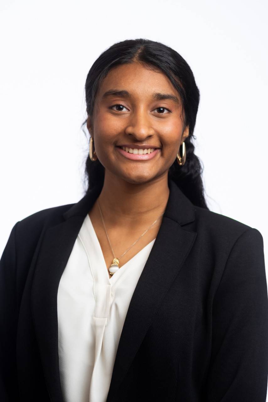 Aparnaa Ananthakrishnan, a young woman wearing a black suit jacket and white blouse, with dark brown hair. 