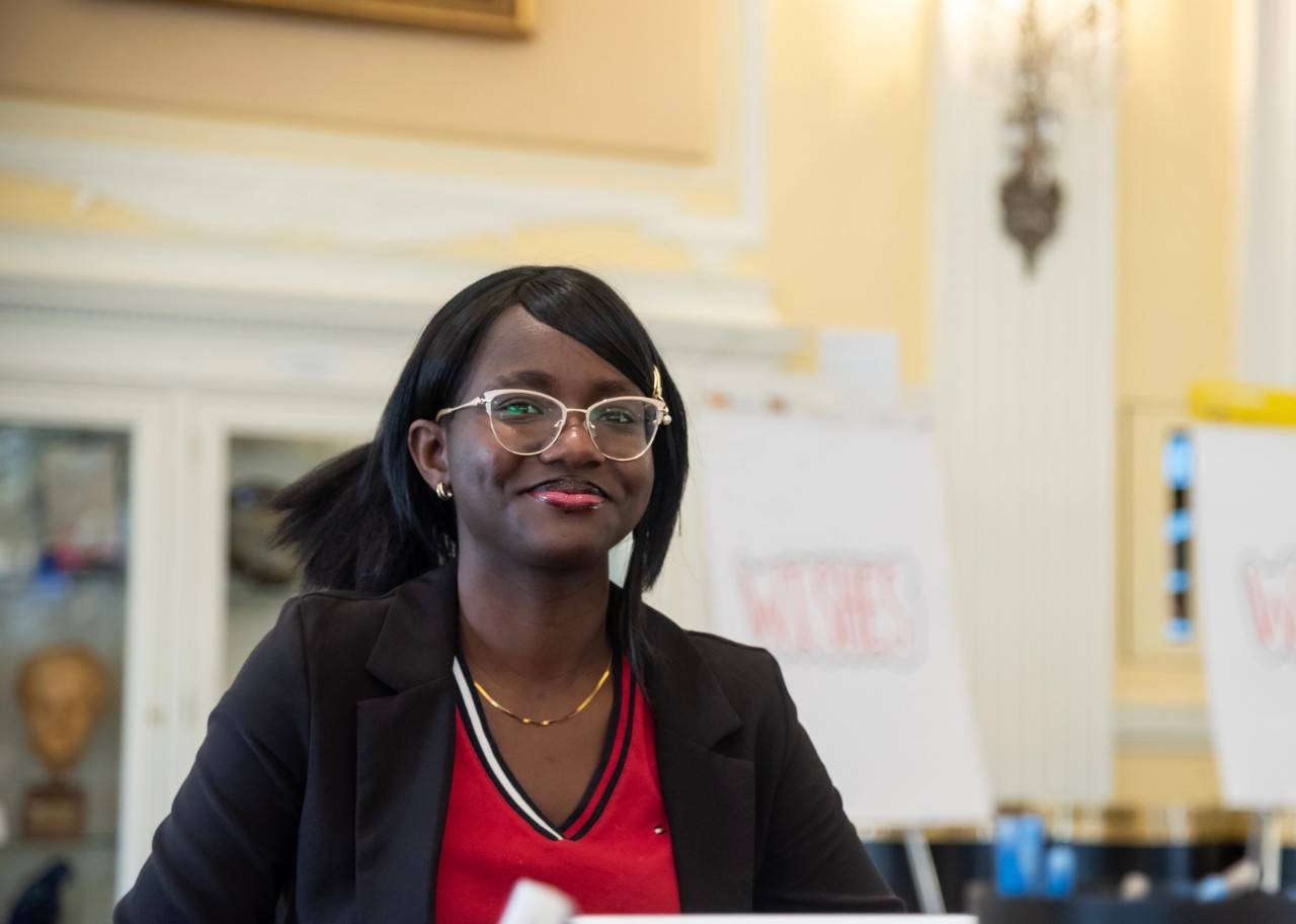 Precious Ikwu,, a young woman wearing a black suit jacket and red blouse, with black hair. 