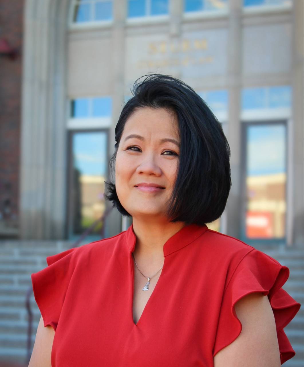 A woman wearing a red shirt with short black hair smiles while posing on the steps of a building.