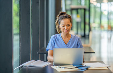 Healthcare professional in scrubs working on a laptop with medical notes and a clipboard, representing secure, time-saving certification through computer adaptive testing.