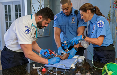 Three EMTs performing pediatric airway management on an infant in a home setting, using a laryngoscope, bag-valve mask, and emergency tools—demonstrating the Practice as You Perform™ Training Model in a real-world critical response.
