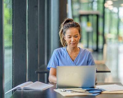 Healthcare professional in scrubs working on a laptop with medical notes and a clipboard, representing secure, time-saving certification through computer adaptive testing.