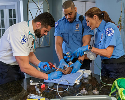 Three EMTs performing pediatric airway management on an infant in a home setting, using a laryngoscope, bag-valve mask, and emergency tools—demonstrating the Practice as You Perform™ Training Model in a real-world critical response.