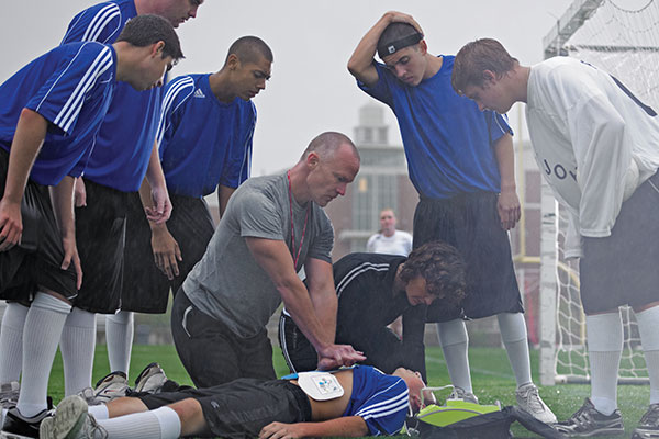 A high school soccer player receives CPR on the field while an AED is applied, with teammates and a coach gathered around during an emergency situation.