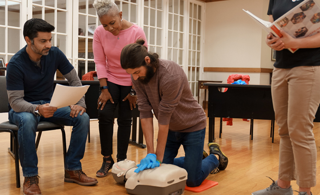 Image of an individual performing CPR on a CPR manikin, onlookers, and a Red Cross Training Provider giving feedback during a group CPR training.