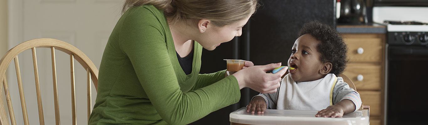 Babysitter feeding a toddler in a high chair.